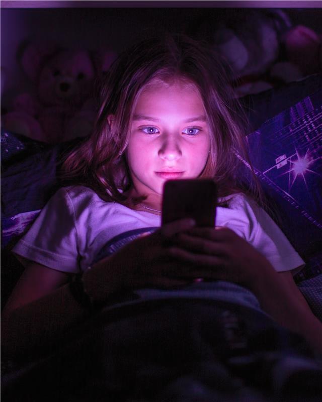 A teenage girl holds a mobile device in bed, her face mesmerized by the glowing screen